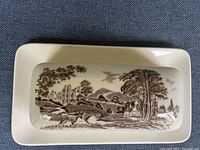 Top view of butter dish lid showing brown rural landscape scene with trees, a stone bridge, castle ruins, animals and houses on cream ceramic base.