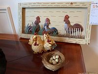 Wide angle showing the framed metal chickens mounted on a wall alongside the ceramic hen statue and nest on a wooden surface