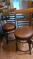 Two bar stools side by side, showing wrought iron frame, wood top slat backrest and round brown faux leather seats.