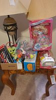 Photo showing wooden table with a variety of toys including boxed Birthday Barbie doll, a plastic container with small toys, and boxed Barbie doll in packaging. Lamp seen in background.