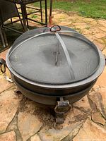 Fire pit viewed from top at an angle, showing the domed mesh cover with handle and circular metal frame.