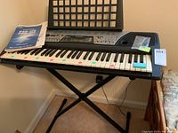Full view of Yamaha PSR-175 keyboard on black X-stand with manual and lesson books placed on the keyboard, showing color-coded keys for learning.