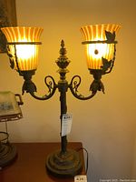 Front and side views of the ornate table lamp with two amber ribbed glass globes and decorative metal leaves on each arm. The lamp is turned on to show illumination through the shades.