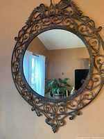 Full view of round mirror with detailed metal frame, showing intricate scrollwork and decorative design at top and bottom of frame, hanging on wall.