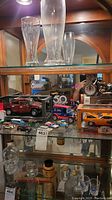 Photo shows a glass display cabinet shelf with a collection of various model cars including a red Hummer H2 SUV with doors open, an orange McLaren MP4-12C in original box, and other collectible miniature vehicles. Beer glasses are visible on the shelf above, and small vintage bottles are below.