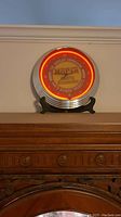 Photo of a round vintage Mopar clock mounted on a black stand, showing the bright orange neon circular light and clear Mopar parts logo.