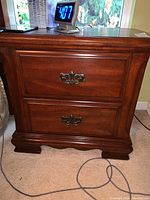 Front view of wooden nightstand with two drawers and decorative metal handles, showing warm brown finish and some scratches.