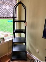 Full view of the black metal pyramid shaped shelf unit with five shelves in descending size, placed by a window with a patterned shade.