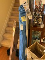 Blue Sport Brella umbrella in its blue storage sleeve with tags attached, standing upright indoors near stairs and wicker baskets.