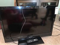 Front view of the Magnavox 32-inch TV with remote on its stand, displaying a black screen and glossy black bezel.