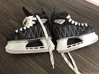 Pair of black kids ice skates sized 13J laced and placed side by side on wooden floor showing the outer sides of both skates with white stitching and white blade holders.