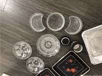Multiple clear glass plates and bowls, two metal containers, and decorative black floral trays displayed on floor.