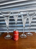 Four tall, clear hand blown glass goblets standing on a wooden surface with a red can for size reference. Goblets have textured stems and smooth flared rims.