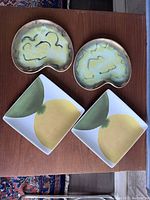 Four decorative plates displayed on a wood surface, showing kidney-shaped glazed ceramic plates and square West Elm plates with a leaf and fruit motif.