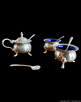 Front and side view of all three miniature silver-plated cellars with cobalt blue glass liners and their matching spoons arranged on black surface.