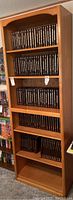 Wood bookcase filled with black DVDs or books, showing all five shelves, four with items and bottom one empty.