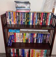 Three wooden shelves with rows of paperback books and a stack of CDs on top shelf.