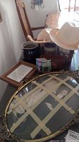 Photo showing an Argus camera in a brown leather case, a straw sun hat, a dark pitcher, a framed note, Sherlock Holmes audiobook CDs, and a vintage oval framed photograph with taped glass damage.