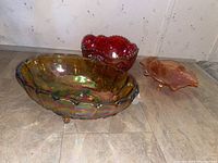 Three carnival glass bowls in different colors positioned on floor against wall - large iridescent bowl in front, medium red bowl in back, small orange-pink bowl to side.