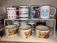 Top rack of assorted coffee mugs including one with red apples, Carnival logo, Raiders logo, and Centennial logo.