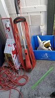 Electric hedge trimmer and weed eater standing upright with orange extension cord coiled on ground, next to packaging box and recycling bin.