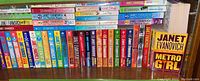 Photo of multiple shelves of Janet Evanovich paperback books showing colorful spines, some co-authored with Charlotte Hughes and Leanne Banks.
