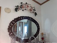 Framed oval wall mirror with dark carved wood frame reflecting room interior and glassware on table beneath it.