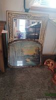 Rectangular mirror with detailed cream and gold frame leaning against wall with vintage print in front of it