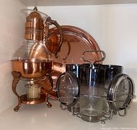 Copper carafe with handle on metal burner stand accompanied by copper serving platter and set of dark-tinted glasses in metal rack.