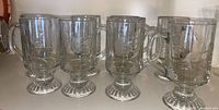Front view of four clear glass coffee mugs showing handles, pedestal bases, and etched vertical line pattern on cups.