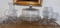 Front view of clear glass pitcher, cake dish with lid, small covered canister, snifter glasses, and glass bowl arranged on wooden surface.