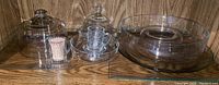 Clear glassware including a round cover, glass tray, coffee mug with saucer, and a covered dish, arranged on wooden surface.