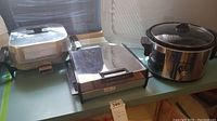 Front view of the lot with all three appliances displayed on a table against a wall with sunlight, showing waffle iron, electric skillet, and crock pot.