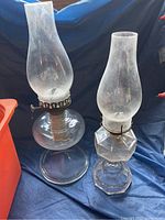 Two antique hurricane oil lamps placed on a blue fabric surface showing their size difference and glass chimney details.