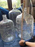 Three clear glass carboys with measuring tape showing heights against natural light window backdrop.