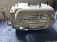Side view of beige plastic cat carrier showing ventilation slots and open metal wire door with latch, placed on dark surface.