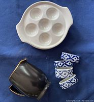 White ceramic escargot plates with seven indentations next to black cream jug and blue and white patterned napkin rings.
