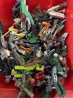 Top-down photo of assorted colorful Bionicle pieces in a red container, showing various limbs, armor, and weapons mixed together.