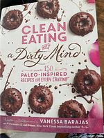 Close-up front cover of 'Clean Eating with a Dirty Mind' cookbook featuring chocolate-covered donuts and pink floral accents.