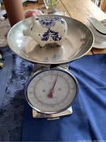 Front view of large mechanical scale with stainless steel bowl containing a decorative white pig-shaped item with blue floral design, dial showing zero mark.