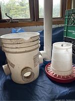 Photo of two white bucket feeders and multiple self chicken feeders including a red base feeder with white feeding ports and a separate cylindrical white feeder.