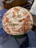 Round paper lantern with a paisley and floral pattern in orange, green, and brown colors on white paper.