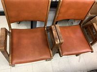 Two MCM vintage chairs with wooden frames, burnt orange vinyl seats and backrests, with armrests, placed side by side.