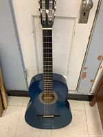 Full view of the small blue classical acoustic guitar standing against a door, showing body, neck, and headstock with six tuning pegs.