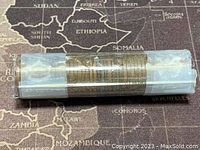 Side view of roll of Canadian one cent coins, showing the coins stacked tightly in a clear plastic tube with white end caps, lying on a vintage map background.