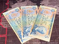 Three Canadian five-dollar banknotes laid out, displaying front side with denomination, crest, and portrait. Banknotes appear slightly worn and have ink markings.