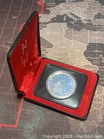 Red velvet-lined display case open showing the coin secured in a clear protective capsule inside a black plastic holder.