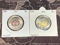 Two Canadian two-dollar coins from 2020 and 2021 in protective holders. The 2020 coin shows a colourful Indigenous mask design. The 2021 coin shows a blue bird on a yellow background.
