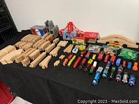 Wide view showing wooden train engines, cars, bridge structures, wooden track pieces, and accessory buildings arranged on a black surface.