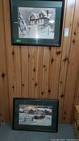 Two framed winter scene prints by Walter Campbell, hanging on wood-paneled wall, one hung and one leaned beneath it.